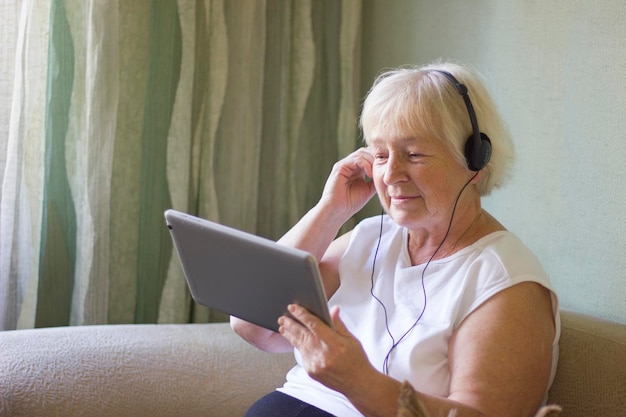 Glimlachende senior vrouw met blond haar maakt een videogesprek thuis met behulp van een digitale tablet en hoofdtelefoon