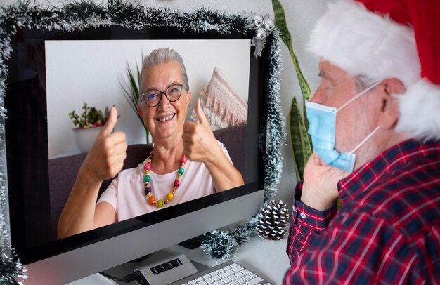 Glimlachende senior vrouw praten over video-oproep voor kerstgroeten met het ontvangen geschenk. Man met kerstmuts draagt chirurgisch masker vanwege quarantaine vanwege coronavirusinfectie