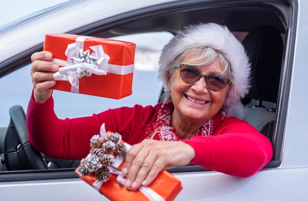 Glimlachende senior vrouw rood gekleed leunt uit het autoraam met een kerstmuts en met kerstcadeautjes