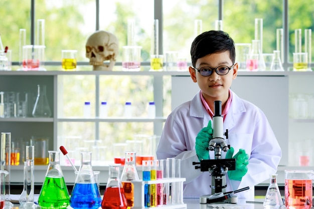 Glimlachende slimme Aziatische jongen in glazen en witte laboratoriumjas staande aan het bureau met microscoop en kolven met kleurrijke vloeistoffen en kijkend naar de camera tijdens scheikundeles in schoollaboratorium.