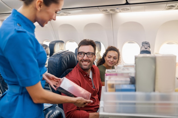 Glimlachende stewardess die producten of geschenken aanbiedt om te kopen aan passagiers in vliegtuigen. Luchtstewardess die met karretje met goederen op gangpad loopt. Reizen, service, transport, vliegtuigconcept