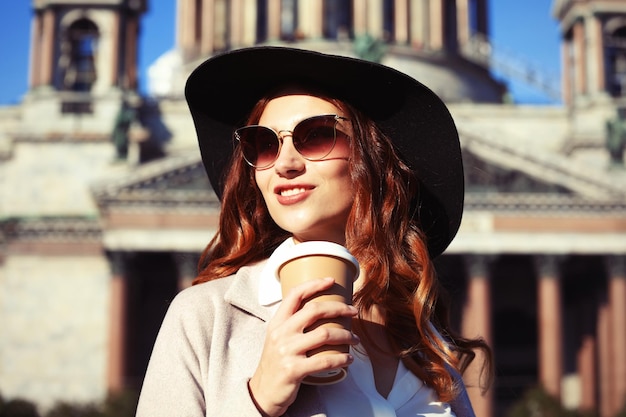 Glimlachende stijlvolle jonge vrouw die koffie drinkt terwijl ze door een stadsstraat loopt