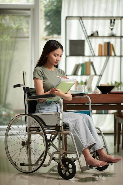 Glimlachende student met een handicap die in een leerboek schrijft terwijl hij in de universiteitsbibliotheek studeert