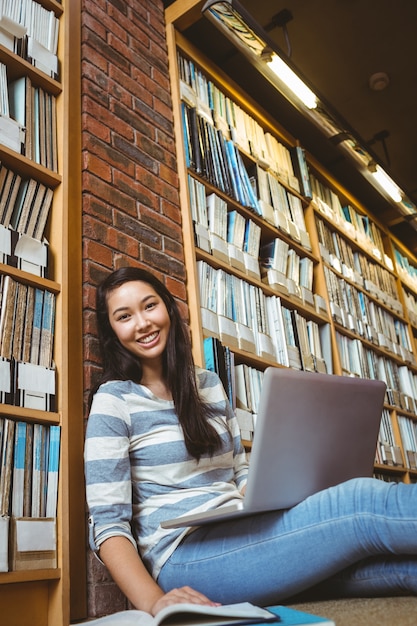 Glimlachende studentenzitting op de vloer tegen muur in bibliotheek die met laptop en boeken bestudeert