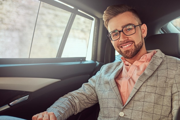 Glimlachende succesvolle stijlvolle jonge zakenman in een grijs pak en roze shirt, rijdend op een achterbank in een luxe auto.
