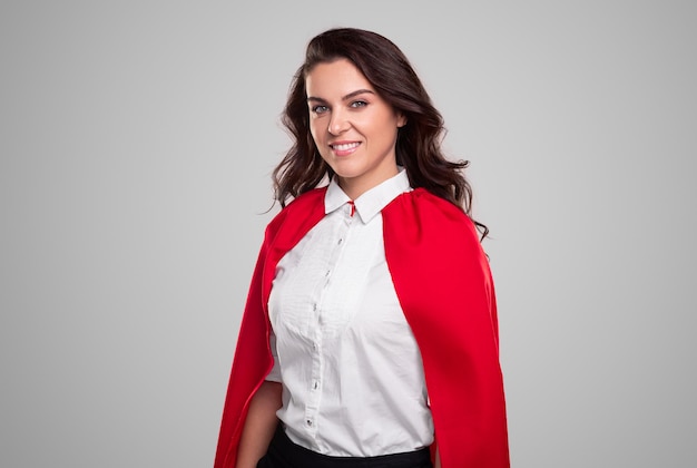 Glimlachende succesvolle volwassen zakenvrouw in rode superheldenmantel die met vertrouwen naar de camera kijkt tegen een grijze achtergrond