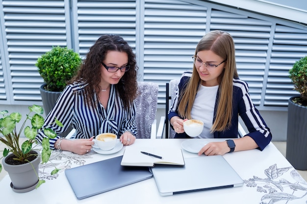 Glimlachende succesvolle vrouwelijke werknemers werken samen, werken op een laptop op het terras, vrouwelijke collega's zijn bezig met het bespreken van ideeën, meisjes in een café tijdens een zakelijke bijeenkomst. bedrijfsconcept