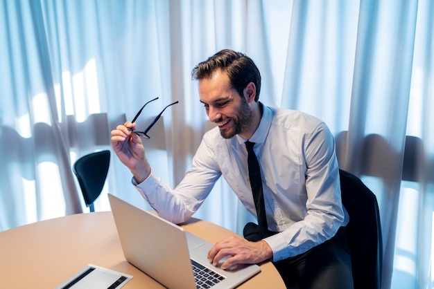 Glimlachende succesvolle zakelijke zakenman met behulp van laptop zittend in zijn kantoor. In handbrillen.