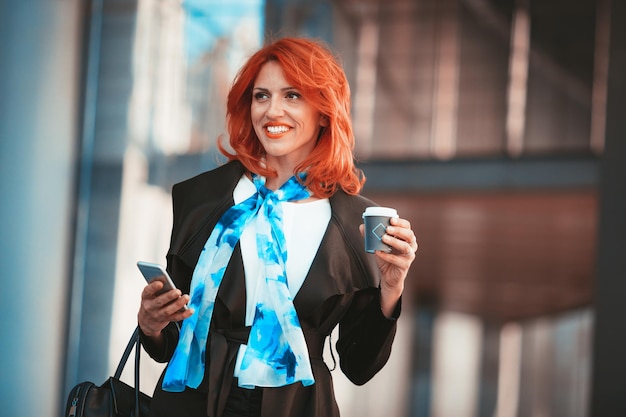 Glimlachende succesvolle zakenvrouw met behulp van smartphone op koffiepauze in office district. Wegkijken.