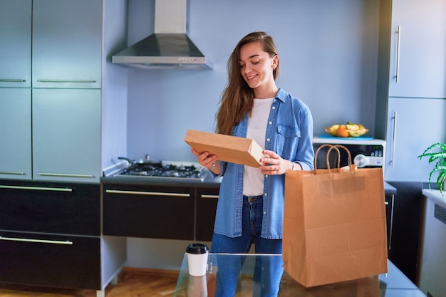 Glimlachende tevreden, gelukkige, volwassen, vrolijke jonge duizendjarige meisjesklant ontving thuis kartonnen zakken met afhaalmaaltijden en drankjes. serviceconcept voor snelle levering