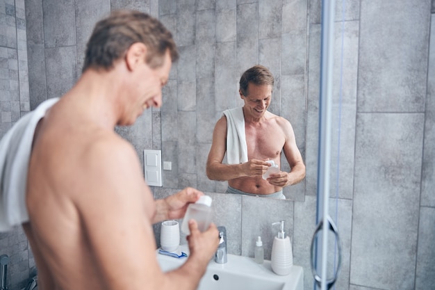 Glimlachende tevreden shirtloze man die naar de eau de cologne in zijn handen kijkt