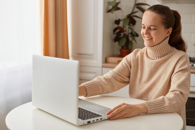 Glimlachende tevreden vrouw met donker haar met een beige trui die in de keuken zit en op een laptop werkt en naar het display kijkt met een vrolijke positieve gezichtsuitdrukking