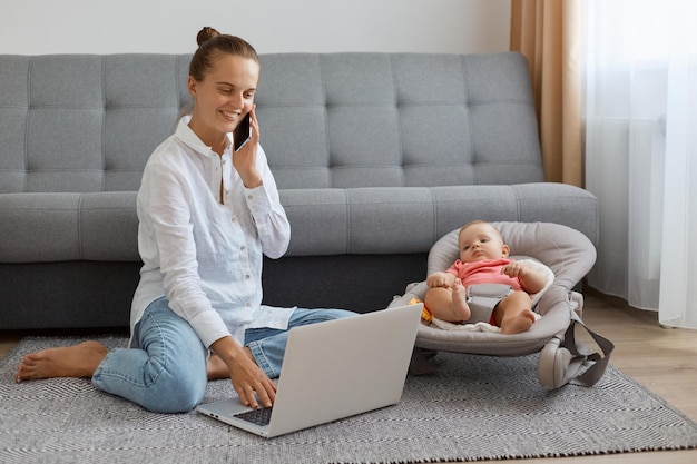 Glimlachende tevreden vrouw met wit overhemd en spijkerbroek zittend op de vloer in de buurt van bank met baby in schommelstoel positieve mama pratende telefoon en werken op draagbare computer