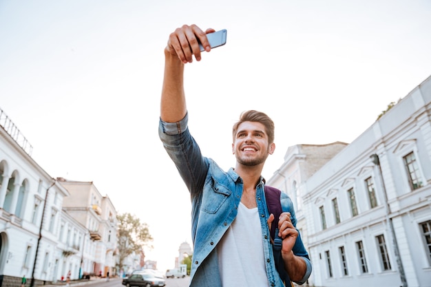 Glimlachende toerist met rugzak die selfies maakt geïsoleerd op prachtige moderne gebouwen in het stadscentrum