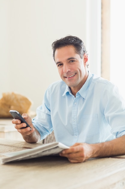Glimlachende toevallige mens met krant en cellphone in keuken