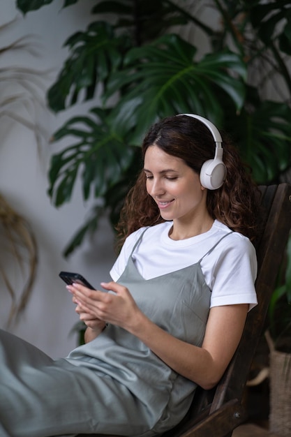 Glimlachende tuinmanvrouw luistert naar muziek, zit in een fauteuil in de binnentuin, ontspannen chatten in smartphone