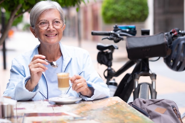 Glimlachende volwassen blanke vrouw zittend aan een tafel buiten met een glas koffie en melk in haar handen Zittend naast haar elektrische fiets