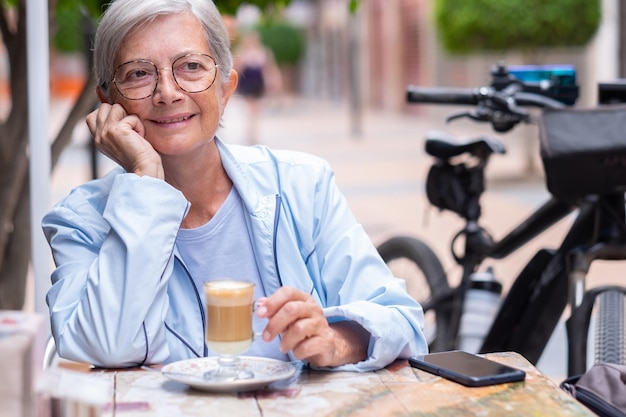 Glimlachende volwassen blanke vrouw zittend aan een tafel buiten met een glas koffie en melk in haar handen Zittend nadenkend in de buurt van haar elektrische fiets