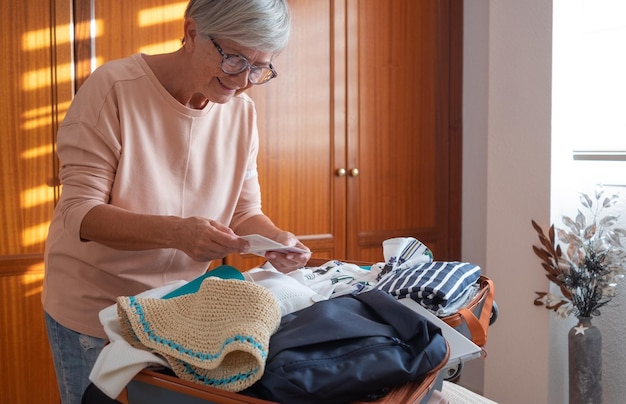 Glimlachende volwassen dame in de slaapkamer controleert haar reisdocumenten terwijl ze haar spullen in een koffer pakt die zich voorbereidt op een reis Reisvoorbereidingsconcept