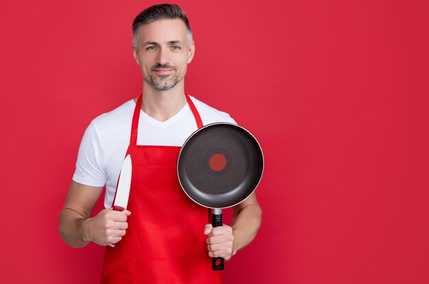 Glimlachende volwassen man chef-kok in schort met koekenpan en mes op rode achtergrond