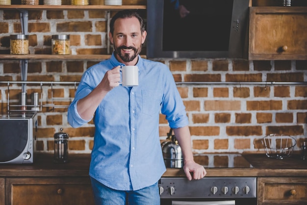 Glimlachende volwassen man met koffiekopje en kijkend naar de camera in de keuken