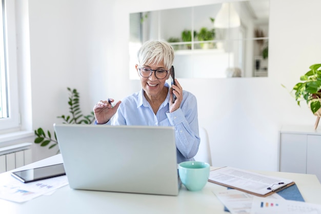 Glimlachende volwassen mooie zakenvrouw met wit haar die op laptop werkt in een helder modern kantoor aan huis. Zakenvrouw die op haar mobiele telefoon praat terwijl ze thuis werkt