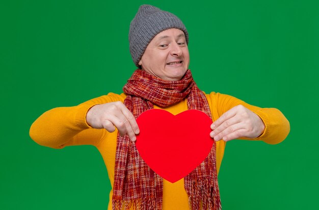 Glimlachende volwassen Slavische man met wintermuts en sjaal om zijn nek die de vorm van een rood hart vasthoudt en bekijkt