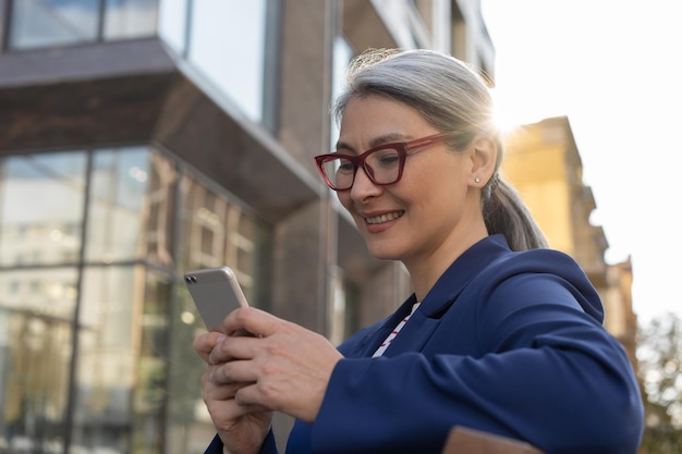 Glimlachende volwassen zakenvrouw die mobiele telefooncommunicatie online gebruikt om e-mail buitenshuis te lezen