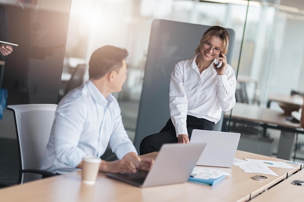 Glimlachende volwassen zakenvrouw zittend op tafel en telefoon praten met cliënt in office