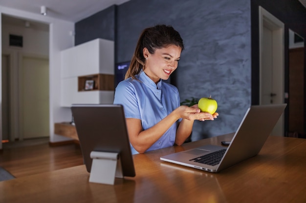 Glimlachende vriendelijke jonge verpleegster om thuis te zitten en appel te houden. Via internet geeft ze adviezen voor een gezond leven.