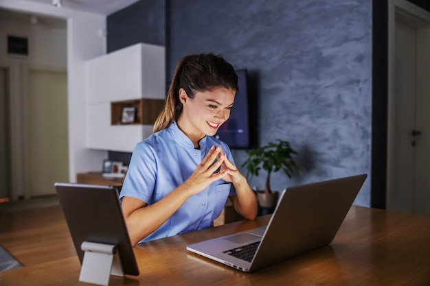 Glimlachende vriendelijke verpleegster om thuis te zitten en adviezen te geven aan de patiënten online. Ze gebruikt tablet en laptop.