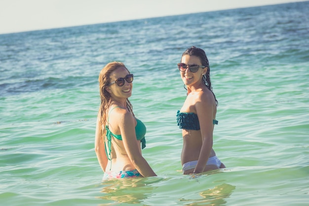 Glimlachende vriendinnen in bikini op turquoise water kijken terug naar de camera op het strand in Thailand