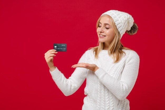 Glimlachende vrij jonge vrouw in witte sweater, hoed die op rood muur achtergrondstudioportret wordt geïsoleerd. Gezonde mode levensstijl, koud seizoen concept. Mock-up kopie ruimte. Wijzende hand op credit bankkaart.