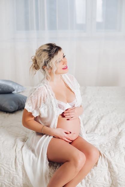 Glimlachende vrij zwangere vrouw in witte lingerie op bed.