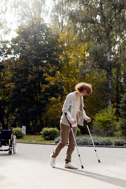 Glimlachende vrolijke man op krukken loopt op straat Een jonge man met een bril herstelt van een blessure op de achtergrond van een rolstoel