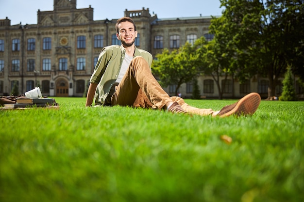 Glimlachende vrolijke, schattige, bebaarde donkerharige mannelijke student in vrijetijdskleding ontspannen op het groene gras