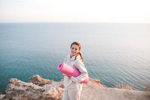 Glimlachende vrolijke vrouw houdt yogamat vast en draagt een trainingshoodie die zich voordeed boven zee