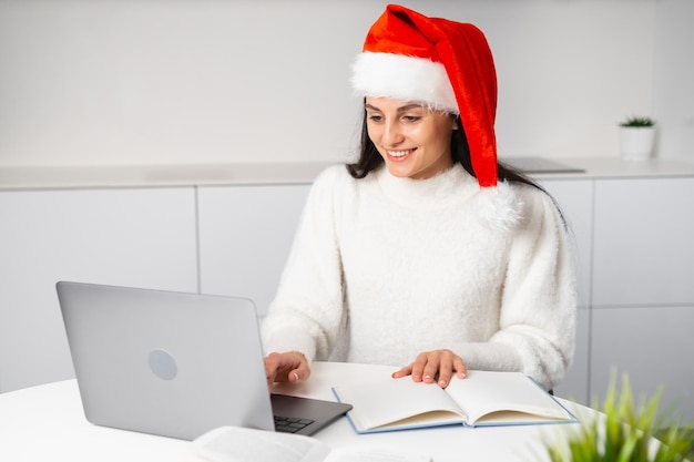 Glimlachende vrouw die de hoed van de Kerstman draagt die laptop voor studie thuis gebruikt