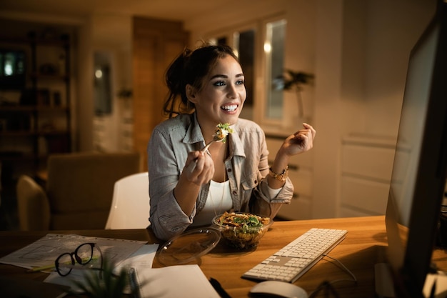 Glimlachende vrouw die een e-mail leest op desktop-pc en 's avonds thuis salade eet