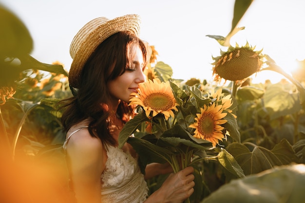 Glimlachende vrouw die een hoed draagt die zich in een veld met zonnebloemen bij zonsondergang bevindt