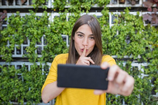 Foto glimlachende vrouw die een selfie maakt op een smartphone met haar vinger op de lippen tegen planten