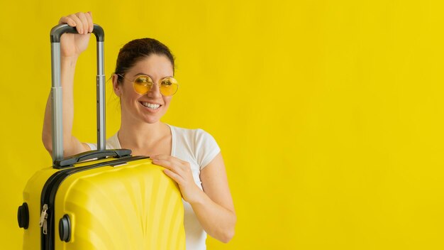 Glimlachende vrouw die in zonnebril koffer met intrekbaar handvat op gele achtergrond houdt