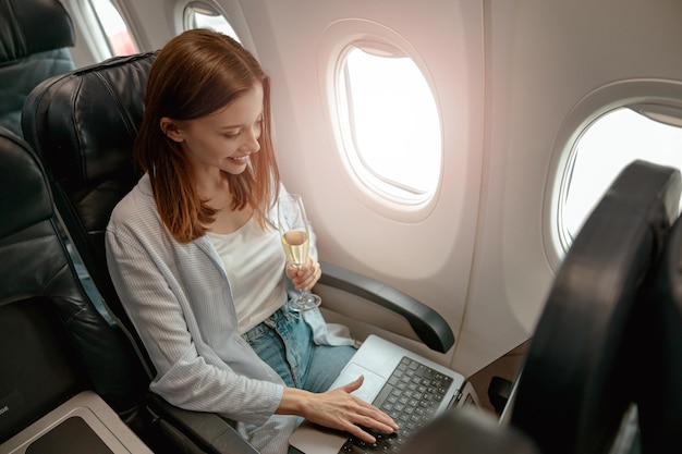 Glimlachende vrouw die laptop gebruikt en champagne drinkt in het vliegtuig