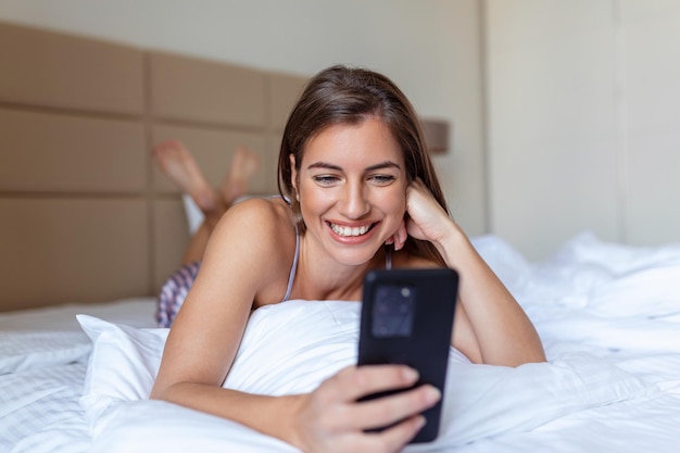 Glimlachende vrouw die mobiele telefoon bekijkt terwijl ze op wit bed ligt