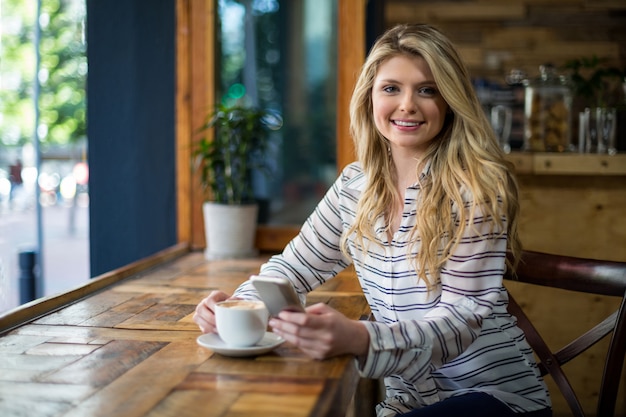 Glimlachende vrouw die mobiele telefoon met behulp van terwijl het hebben van koffie in koffie