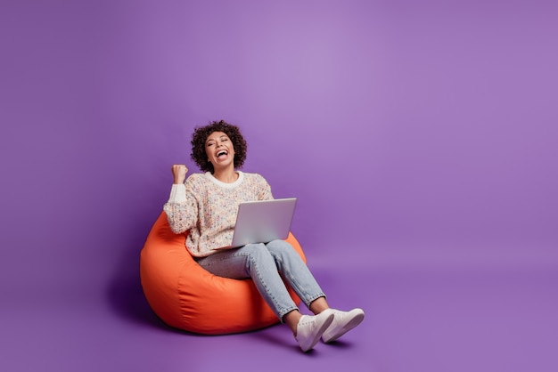 Glimlachende vrouw die op laptopcomputer typt, verheugt zich over de overwinning, hef de vuist op, zit een zitzak op de paarse muur