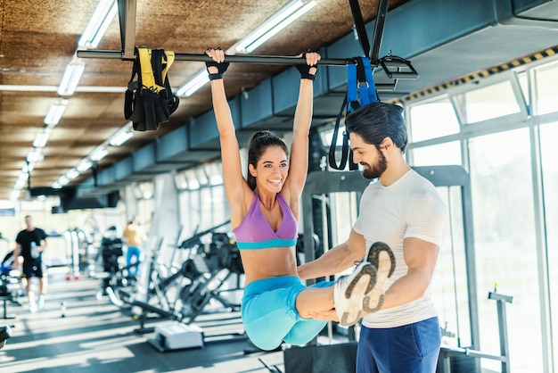 Glimlachende vrouw die uithoudingsvermogen op staaf doet terwijl haar persoonlijke trainer haar helpt. gym interieur.