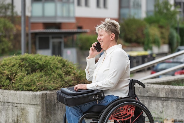 Foto glimlachende vrouw een rolstoel gebruiker met een laptop tas in haar schoot met behulp van een smartphone om te praten handicap