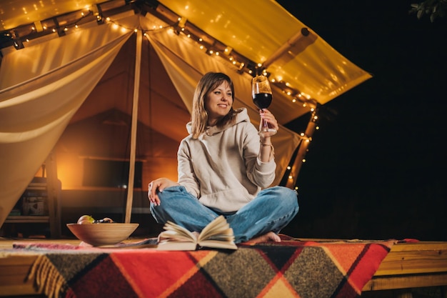 Glimlachende vrouw freelancer wijn drinken en boek lezen zittend in gezellige glamping tent in herfst avond Luxe camping tent voor buiten vakantie en vakantie Lifestyle concept