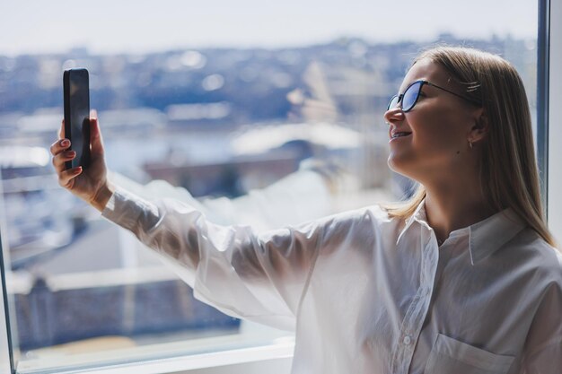 Glimlachende vrouw in bril in vrijetijdskleding kijken naar een positieve video op een telefoon verbonden met wifi een vrouwelijke blogger in een wit overhemd tegen een groot raam genieten van ontspanning tijdens werken op afstand